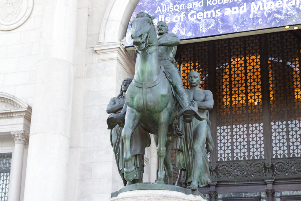 People enter the American Museum of Natural History in 2021 past a statue of Theodore Roosevelt on horseback with a Native American man on one side and an African man on the other at New York City.