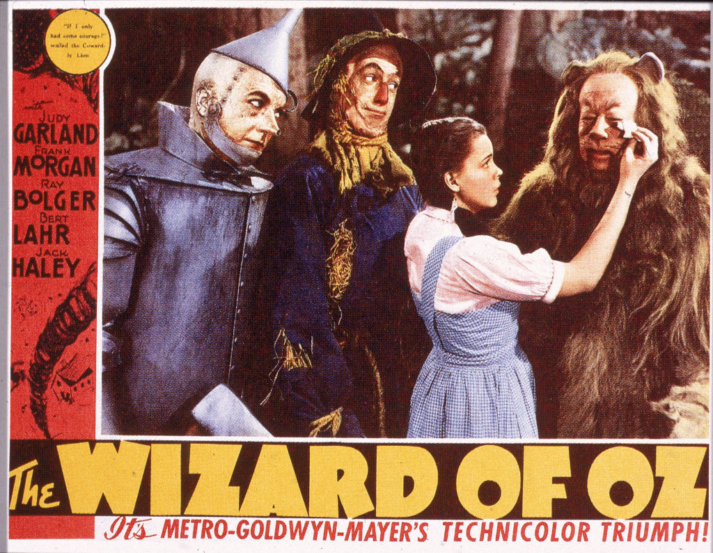 A lobby card from the film 'The Wizard Of Oz,' shows a film still of a scene in which American actress Judy Garland (1922 - 1969) (as Dorothy) wipes tears from the eyes of actor Bert Lahr (1895 - 1967) (as the Cowardly Lion), while watched by Jack Haley (1898 - 1979) (as the Tin Man) (left), and Ray Bolger (1904 - 1987) (as the Scarecrow), 1939. The film was directed by Victor Fleming.