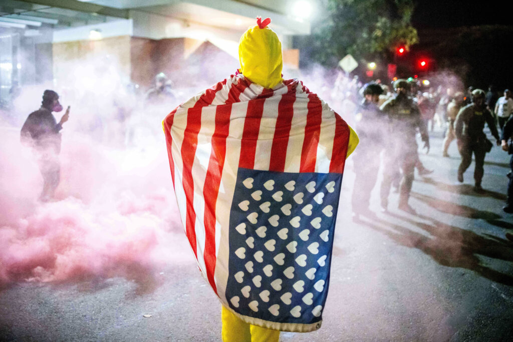 A protester, draped in an altered American flag, watches as law enforcement officers try to disperse protesters near a U.S. Immigration and Customs Enforcement facility in Portland, Ore. on Sunday, Oct. 5, 2025.